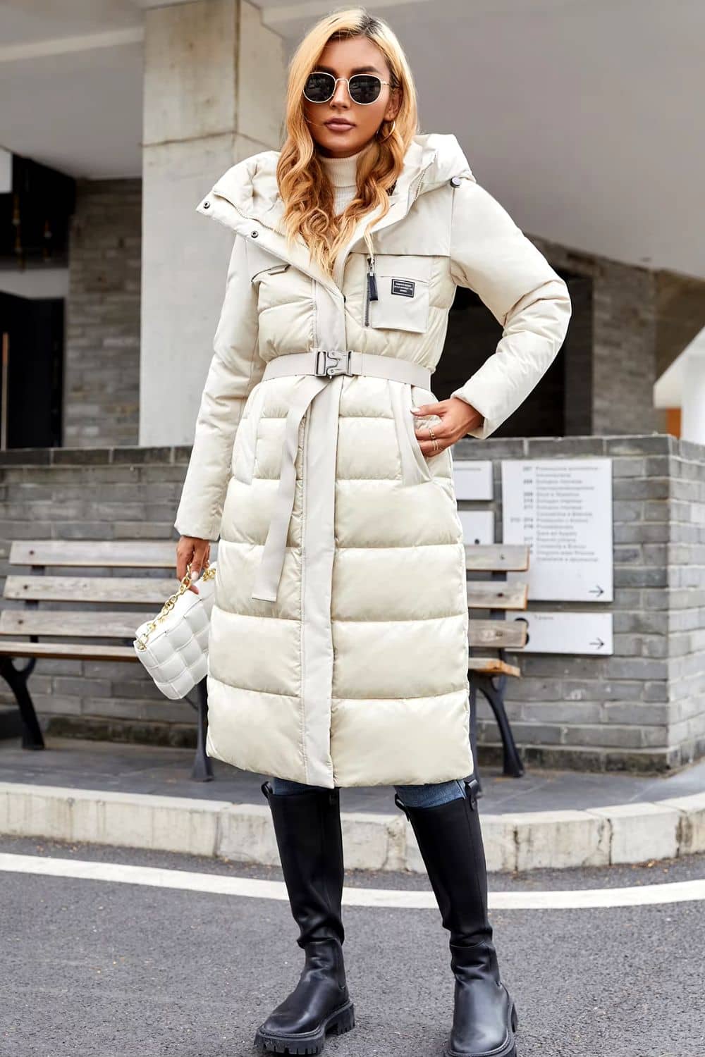A woman wearing a beige quilted puffer jacket with a belt, hood with drawcord, and black boots standing outdoors.