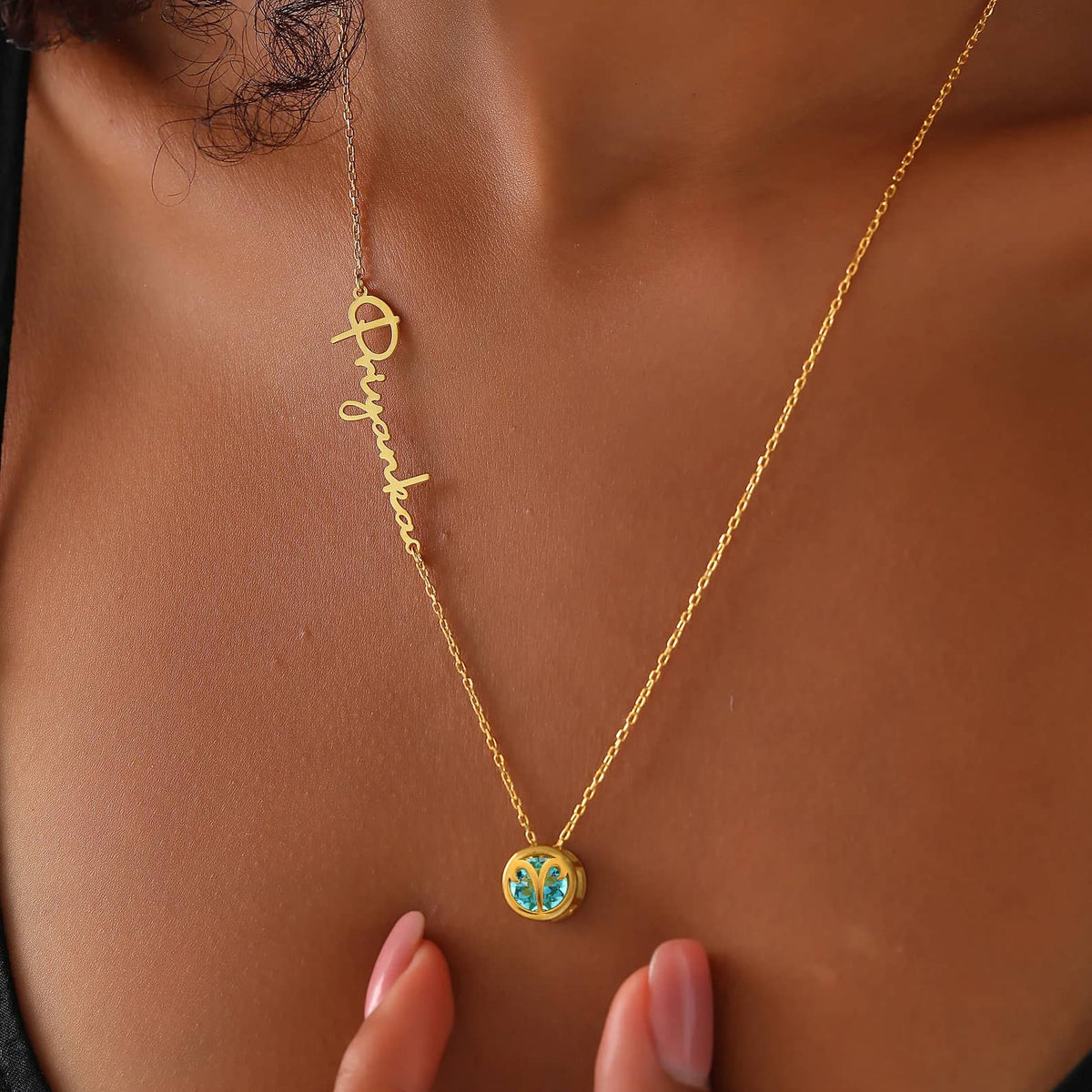 A close-up image of a custom birthstone necklace with a gold cable chain and a round Aries pendant with an aquamarine gemstone embedded, and engraved with a name.