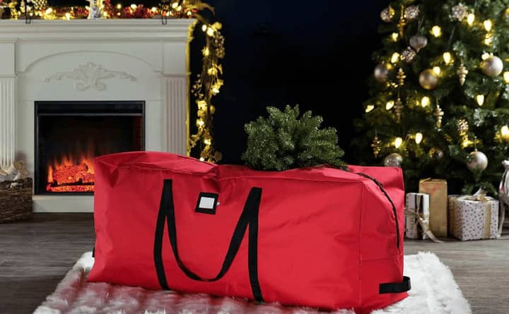 A red Christmas tree storage bag with handles, placed against the backdrop of a decorated Christmas tree and a fireplace.