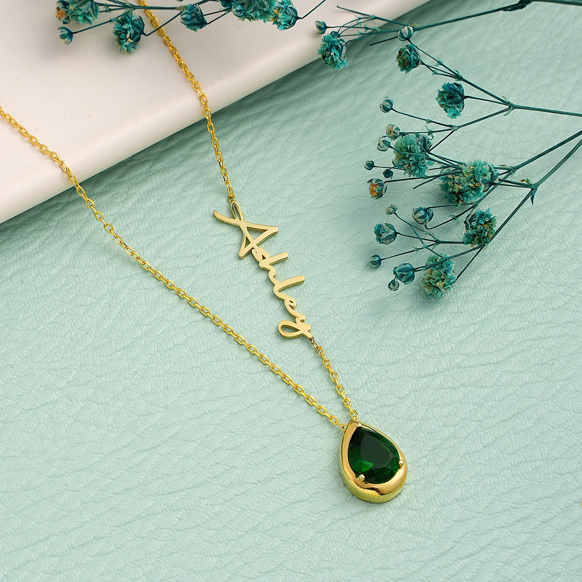 Gold necklace with an emerald teardrop pendant on a light green surface with dried flowers.