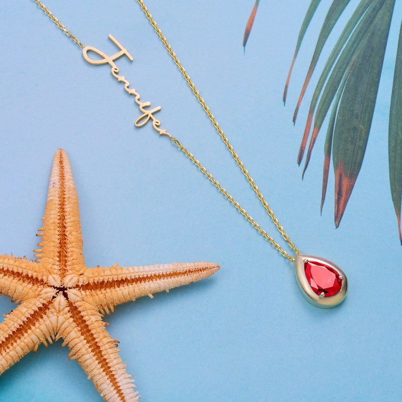 A gold pendant necklace with a teardrop garnet birthstone and a name engraved on the chain.