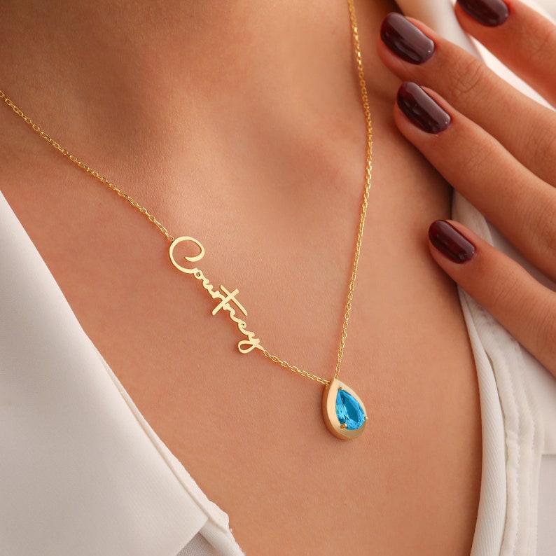 A woman wearing a gold vermeil custom name necklace with a teardrop aquamarine birthstone and personalized engraving on the chain.