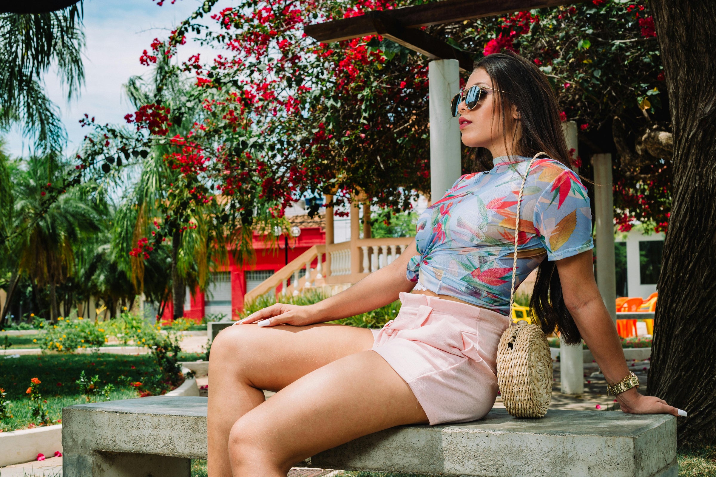 Woman sitting on a bench in a garden with colorful floral patterned shirt and pink shorts.