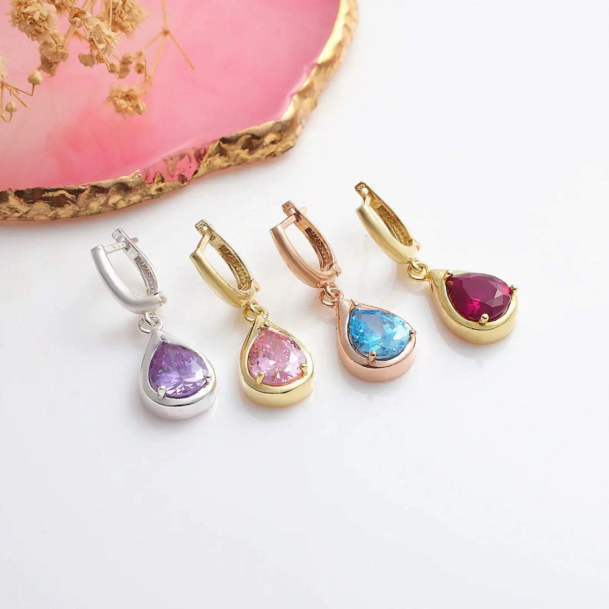 A set of teardrop-shaped birthstone earrings in various colors, displayed alongside a pink agate slice on a white surface.