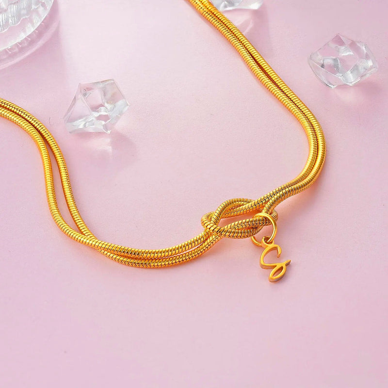 Double gold snake chain necklace tied in love knot with dangling initial charm on pink background