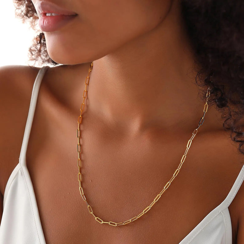 A close-up of a woman wearing a delicate gold chain necklace with elongated oval links, styled with a white spaghetti-strap top.